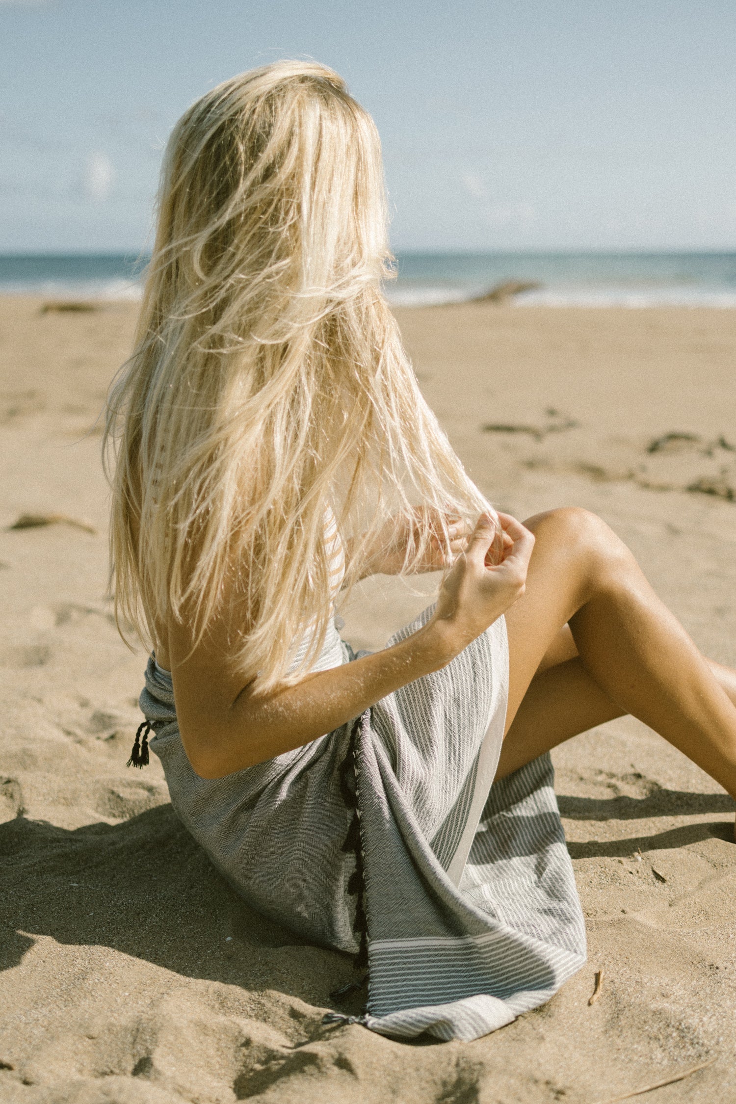 A person sitting on a sandy beach with a grey striped towel wrapped around them, facing away from the camera with long blonde hair visible