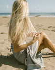 A person sitting on a sandy beach with a grey striped towel wrapped around them, facing away from the camera with long blonde hair visible