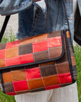 A person holding a crossbody bag with a patchwork design made from upcycled leather, featuring a black nylon strap and interior pockets, in brown, red, and blue colors.