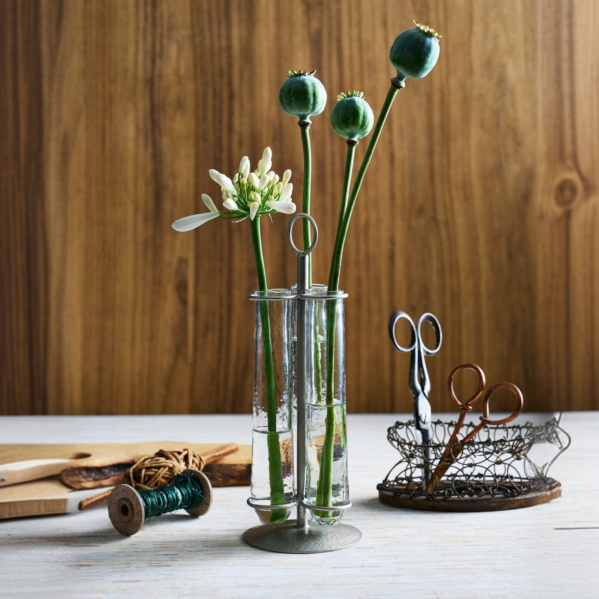 A table vase with three glass stems, featuring a hammered texture and pewter finish, displayed on a wooden surface with a pair of scissors and decorative items in the background.