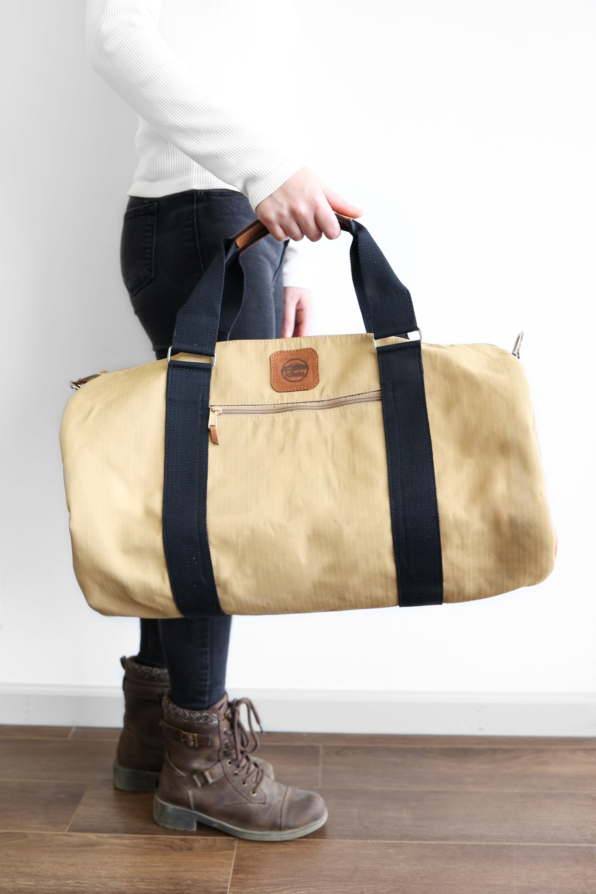 A person holding a beige and brown duffel bag with black straps and a tan leather bottom.