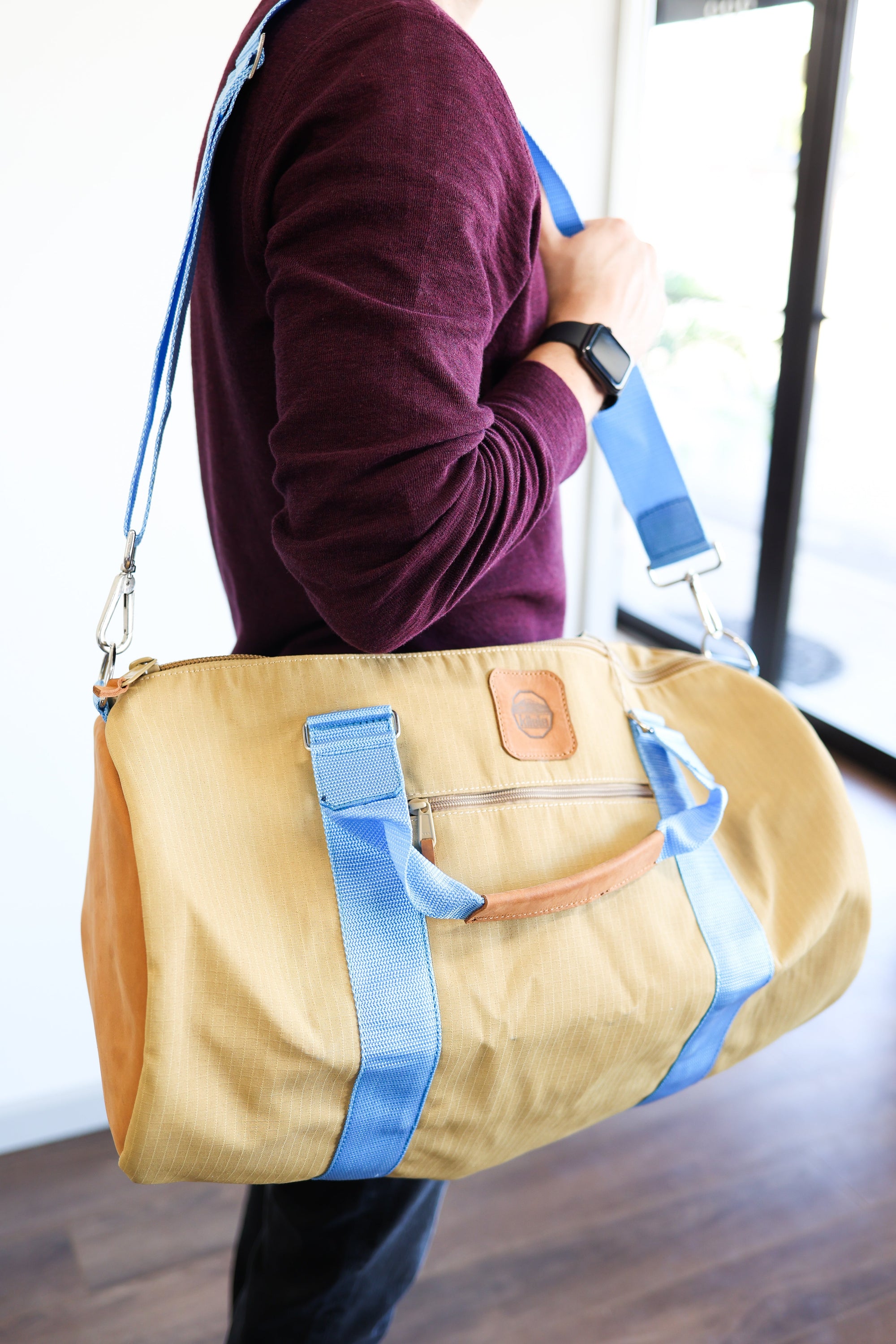 A person holding a beige and blue duffel bag with black straps and a tan leather bottom with the strap around their shoulder.