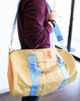 A person holding a beige and blue duffel bag with black straps and a tan leather bottom with the strap around their shoulder.