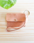 Small pink leather pouch with a strap on a light wooden surface with green leaves in the corner.