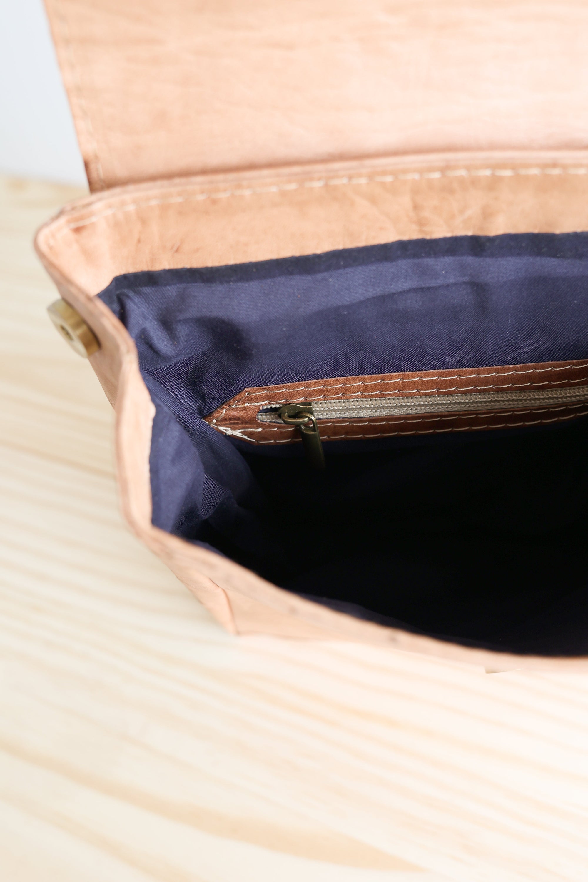 Close-up of a beige leather bag with a navy interior on a light wooden surface.