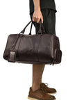 A person carrying a brown leather weekender bag with a leather handle and visible stitching details.