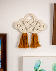 Decorative cotton wall art with braided design and tassels on a wall next to framed pictures.