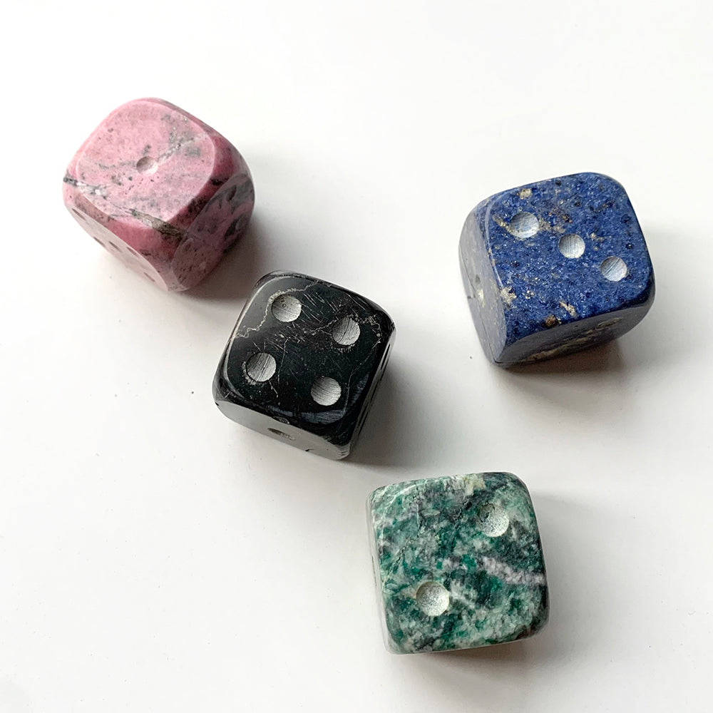 A set of four hand-carved gemstone dice, in pink, blue, black, and green colors, displayed on a white surface.