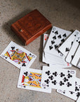 A closed embossed leather box in brown color with intricate floral patterns on the cover alongside the playing cards that go inside of it on top of a table.
