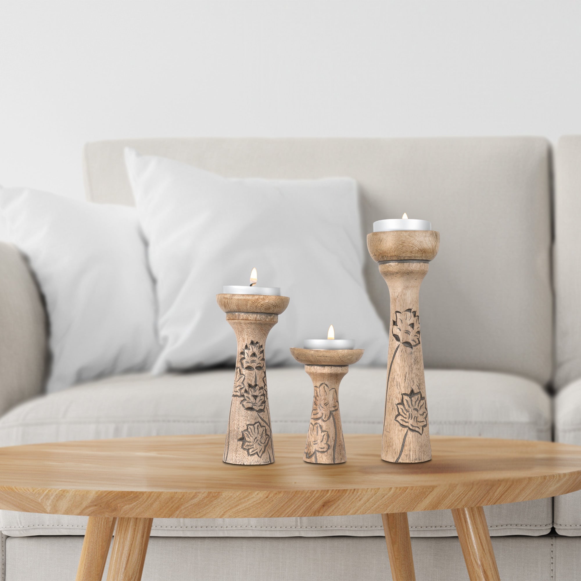 A set of three wooden pillar candleholders with lotus vine carvings, in varying sizes on top of a table holding lit soy wax candles with a couch in the background