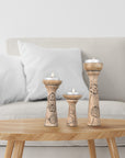A set of three wooden pillar candleholders with lotus vine carvings, in varying sizes on top of a table holding lit soy wax candles with a couch in the background