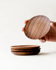 A person holding a round teak wooden coaster with three additional coasters placed together on a table