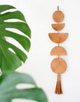 Decorative wall hanging with geometric moon shapes and tassels next to a green leaf on a white background