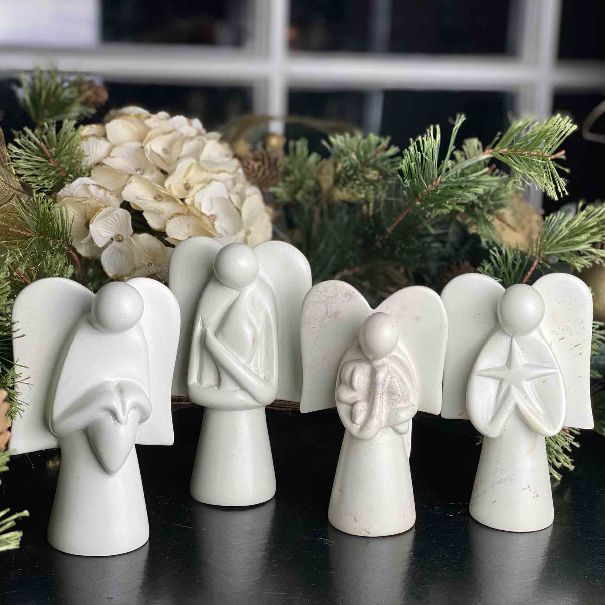 Four handmade stone sculptures of angels holding stars in front of a christmas tree