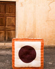 A square-shaped floor cushion with a brown and beige color scheme, featuring a hand-pressed circular design, placed against an old wooden door.