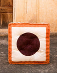A square-shaped floor cushion with a brown and beige color scheme, featuring a hand-pressed circular design, placed against an old wooden door.