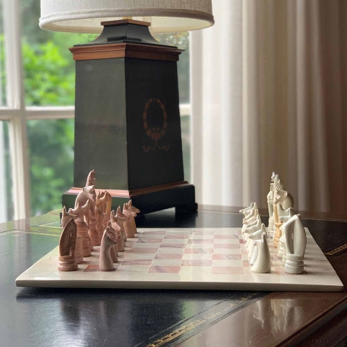 A hand-carved soapstone chess set with a green and pink checkered board and beige, pink, and white stone pieces zoomed in on the white pieces on a table near a lamp.