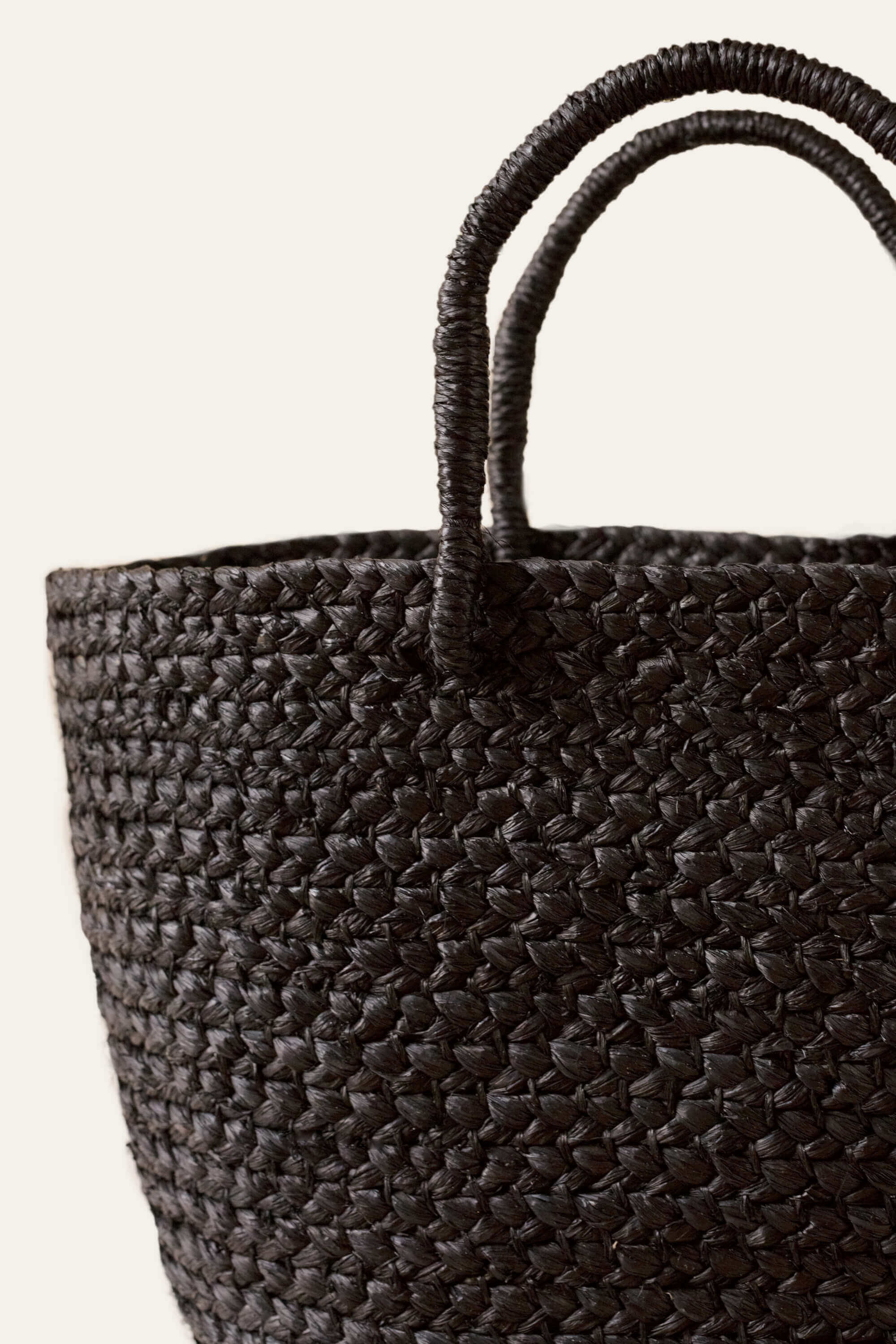 An up close photo of a black braided raffia day bag with sturdy handles and a unlined interior displayed on a white background.