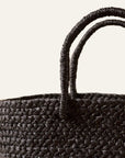 An up close photo of a black braided raffia day bag with sturdy handles and a unlined interior displayed on a white background.