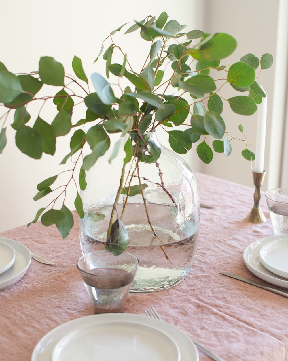 Clear vase with green leaves on a table setting with plates and cutlery.