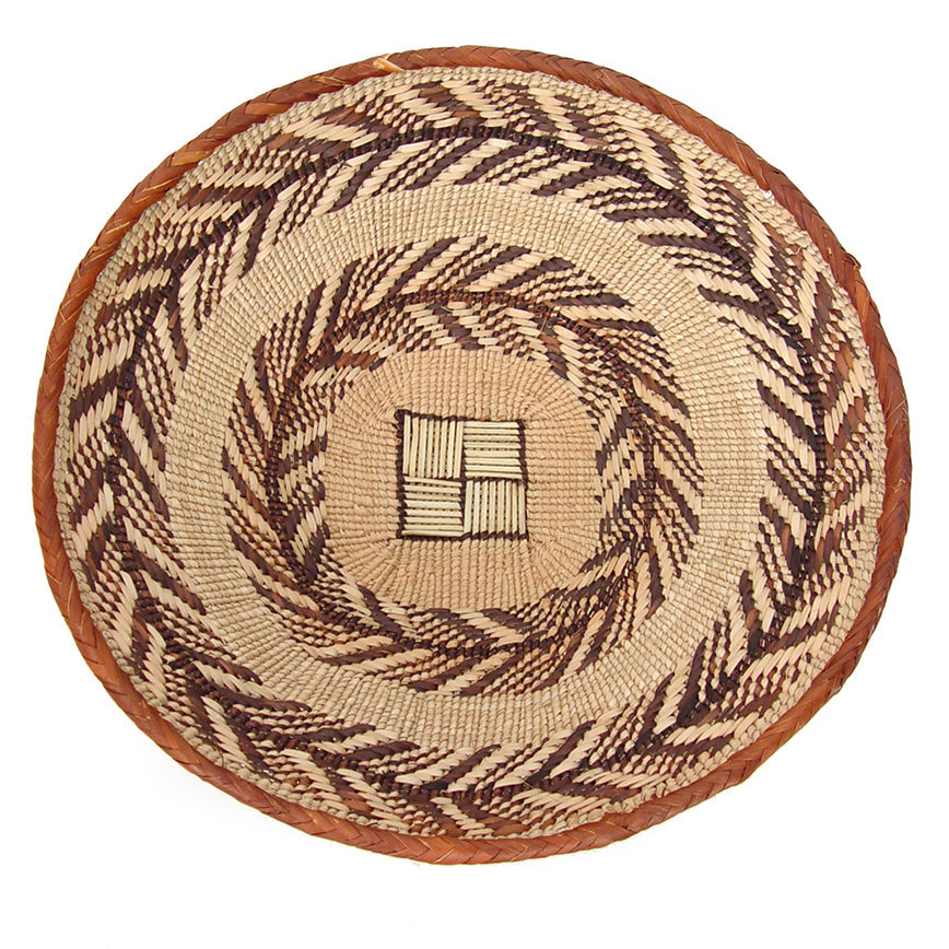 A small, round, handcrafted basket with a geometric pattern, predominantly in beige with brown accents, made by the Tonga people of Zimbabwe