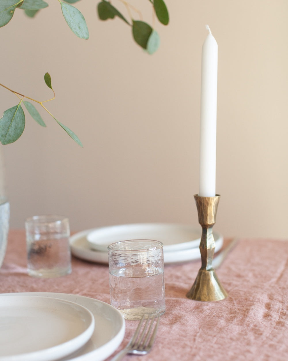 Table setting with plates, glasses, a candle in a brass holder, and greenery on a neutral background.