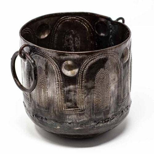 A handcrafted hammered metal container with round handles, made from recycled steel, with a darkened patina finish
