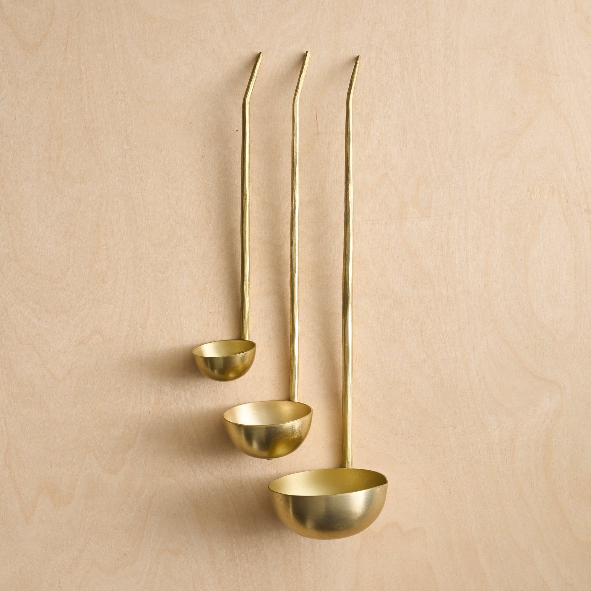 A set of three gold-colored brass ladles of assorted sizes with hammered handles, displayed against a wooden background