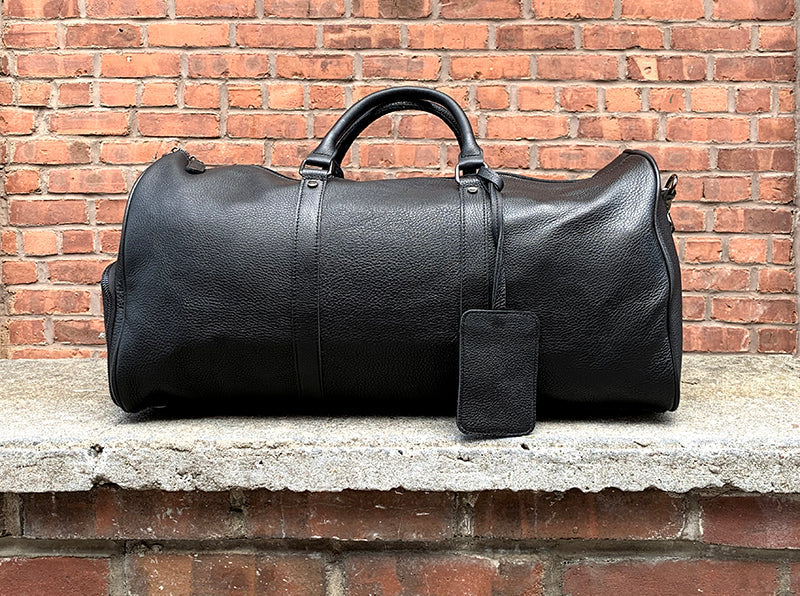 a black leather weekender bag with a leather handle and visible stitching details on a ledge with a brick background.