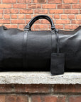 a black leather weekender bag with a leather handle and visible stitching details on a ledge with a brick background.