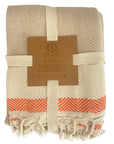 A beige throw blanket with orange stripes and hand-loomed details, displayed with its packaging