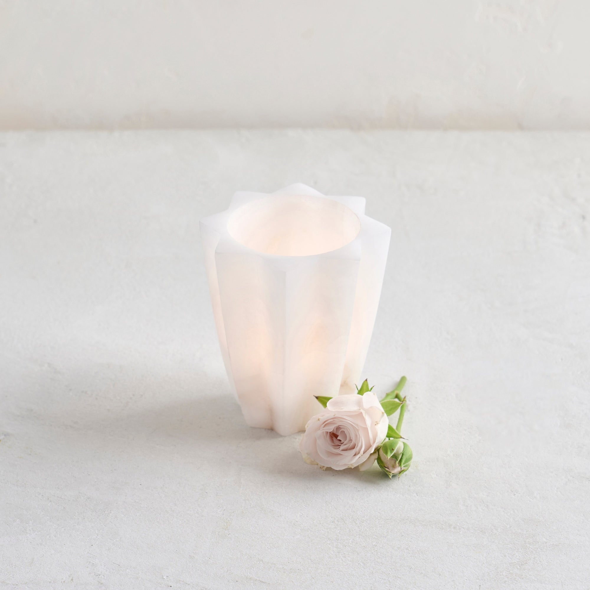 A handcrafted white alabaster votive cup with a star-shaped design, displayed next to a small pink flower.