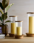 Three glass candle holders with wooden bases on a table, one with a plant and books in the background.