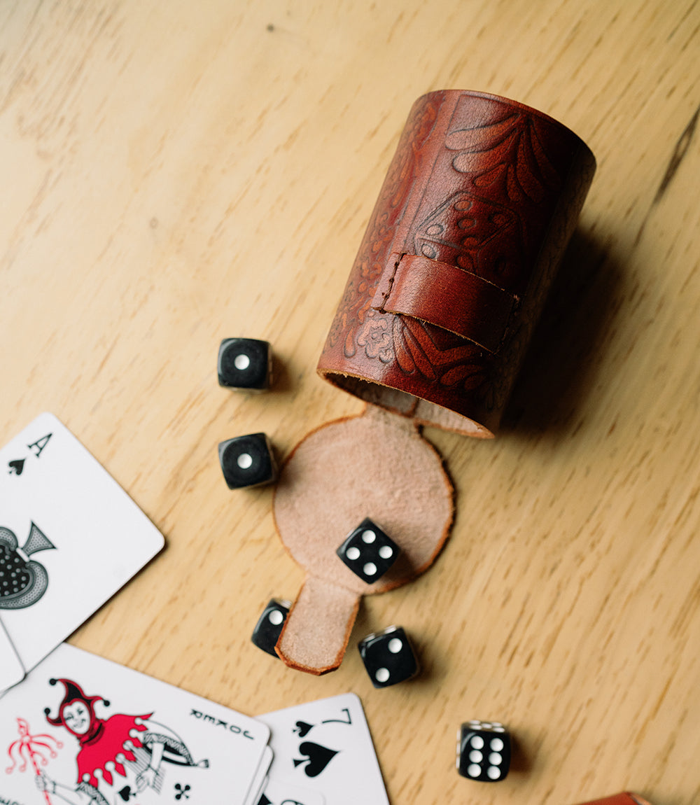 A hand tooled brown leather dice cup open on its side with the dice out next to cards on a wooden table.