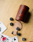 A hand tooled brown leather dice cup open on its side with the dice out next to cards on a wooden table.