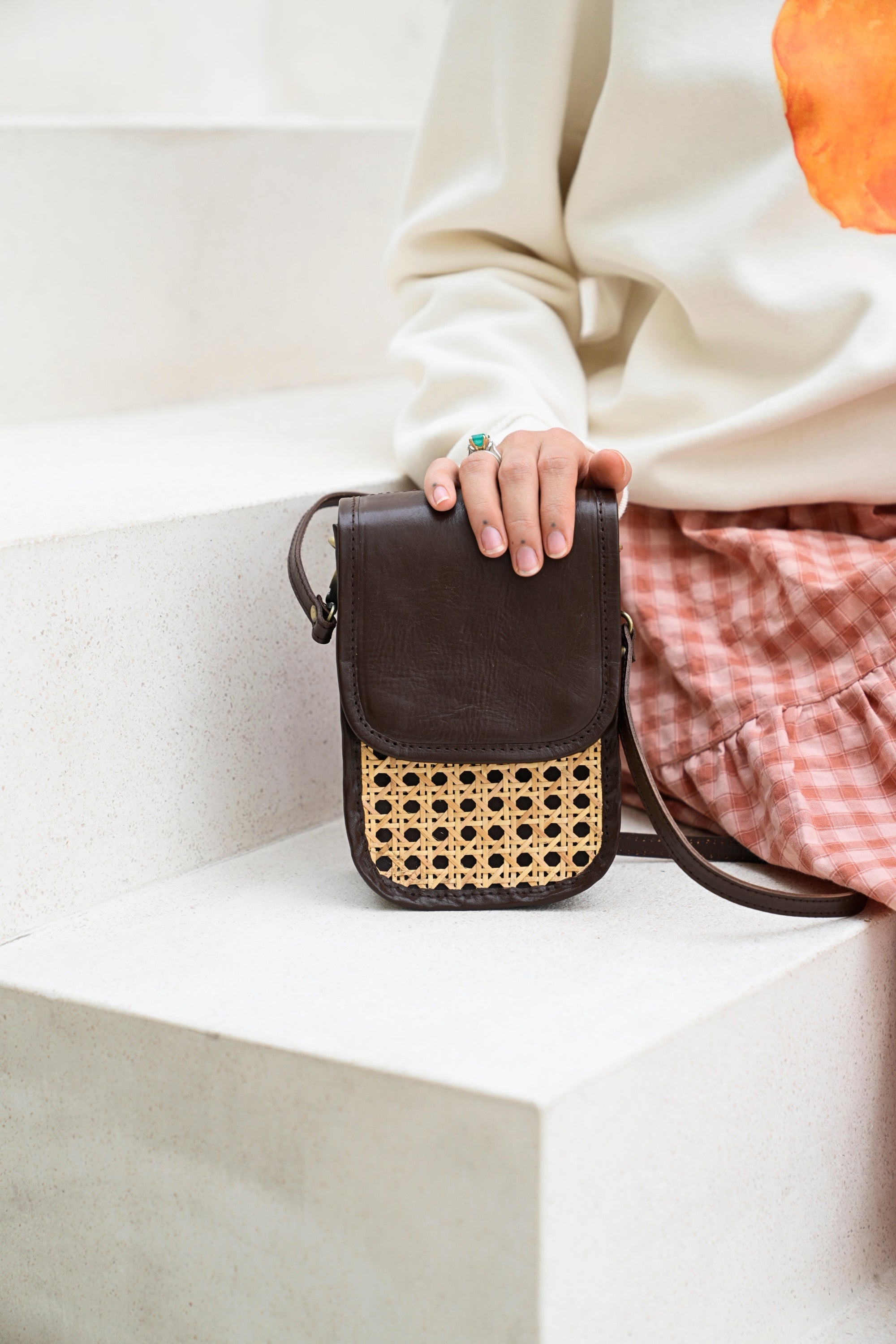 A model with her hand on a brown clutch crossbody bag with rattan cane pattern on the front and leather trim, featuring a detachable shoulder strap.