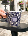 A hand holding mexican ceramic mug with blue floral patterns outside in a backyard on a black table