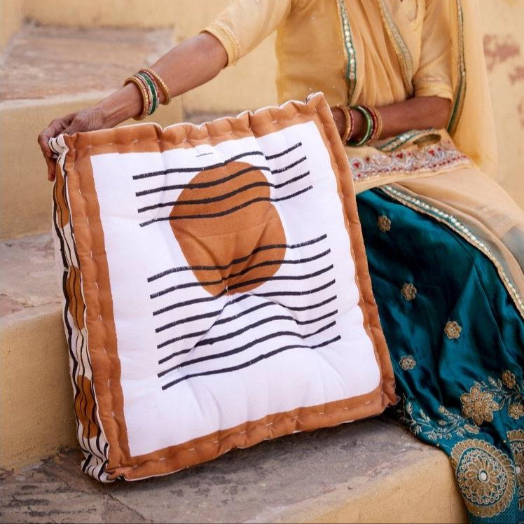 A handcrafted square floor cushion with a geometric pattern, predominantly white with a large brown circle and black stripes, is displayed in the lap of a person dressed in traditional attire