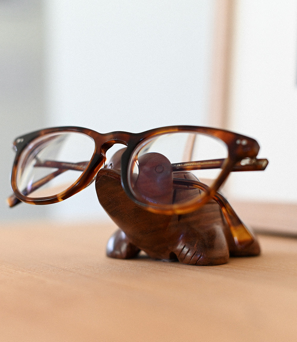 A turtle-shaped wooden glasses holder with a pair of eyeglasses resting on top of it on top of a wooden desk