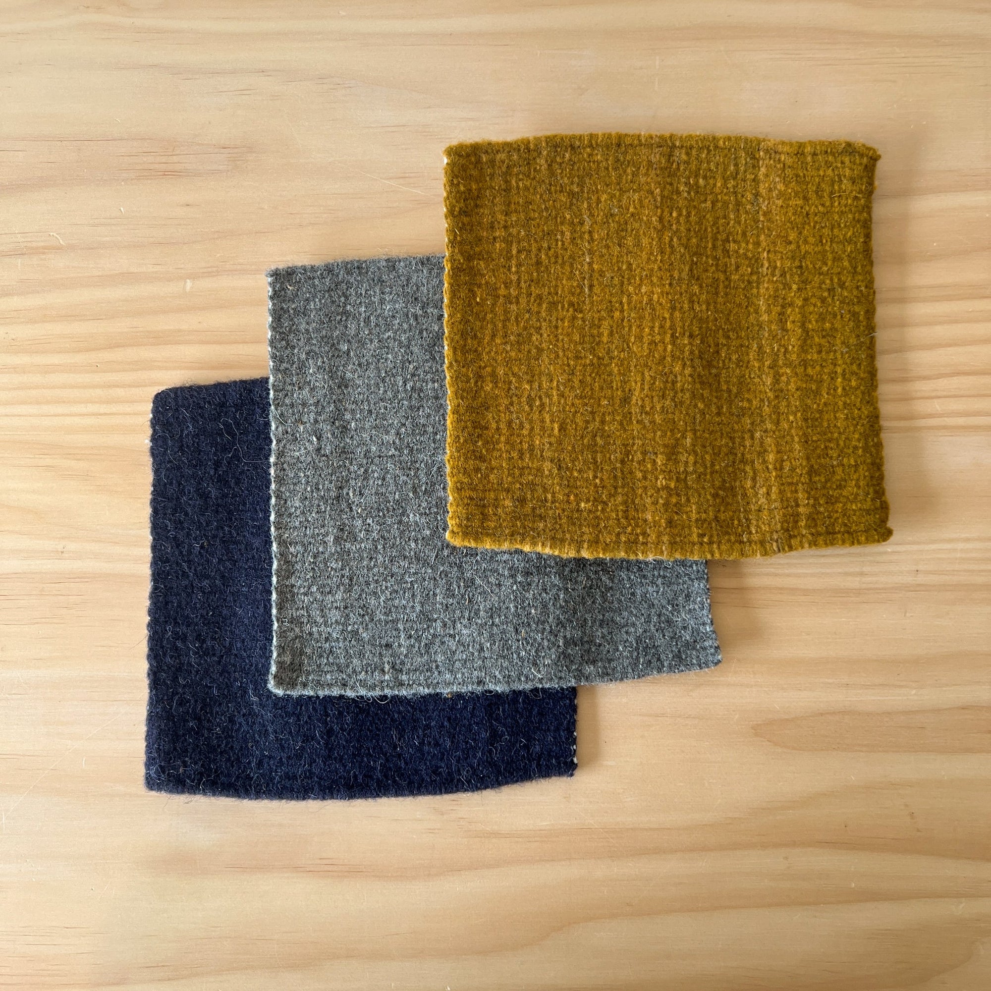 Three wool trivets in blue, gray, and mustard yellow on a wooden surface