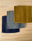 Three wool trivets in blue, gray, and mustard yellow on a wooden surface