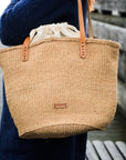 Person holding a woven tote bag with leather straps outdoors.