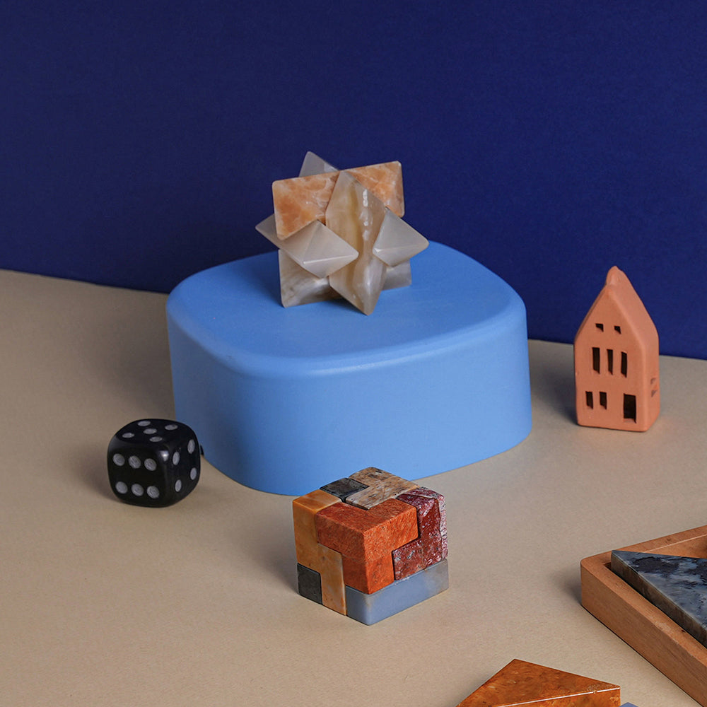 A creamy onyx burr star puzzle, with a complex star shape made from interlocking notched sticks on a blue table next to dice and a rubix cube.