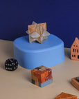 A creamy onyx burr star puzzle, with a complex star shape made from interlocking notched sticks on a blue table next to dice and a rubix cube.