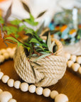 Decorative plant pot wrapped in twine on a wooden surface with beads and a blurred background