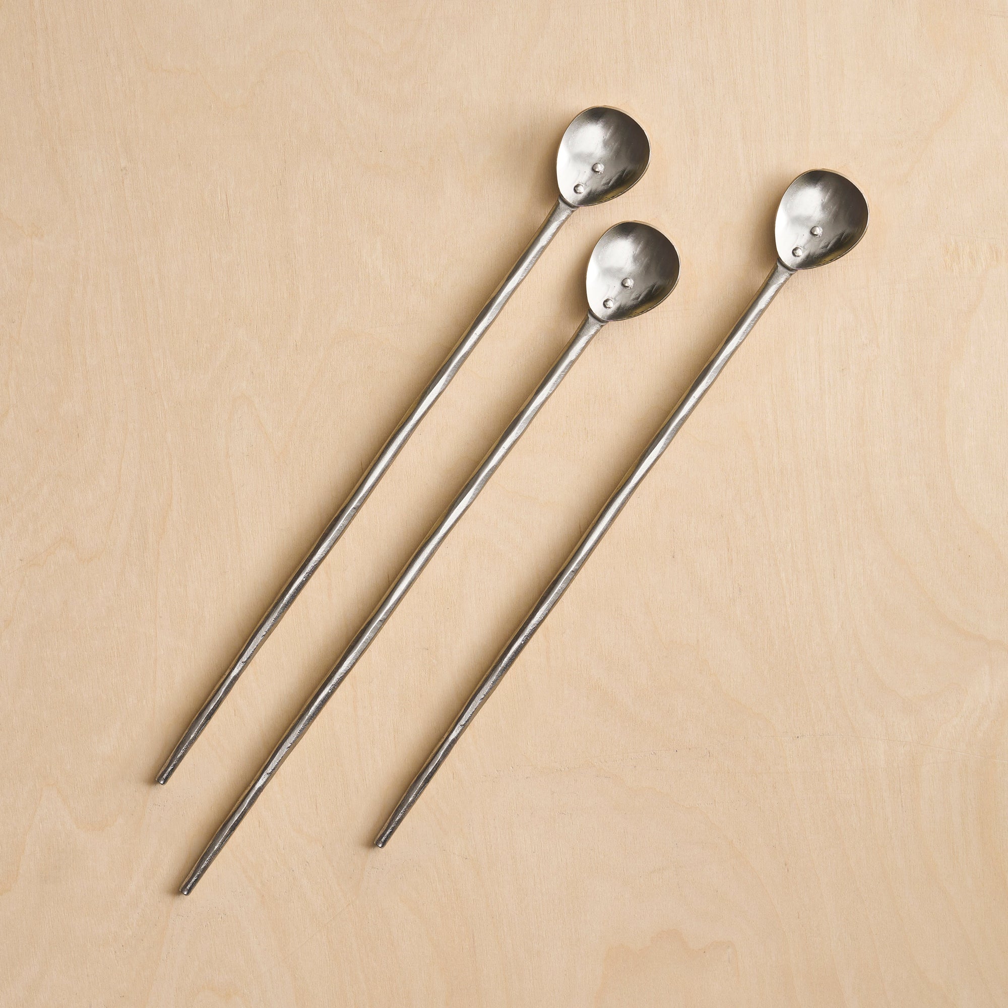 A set of three pewter tasting spoons with long handles, laid out on a wooden surface