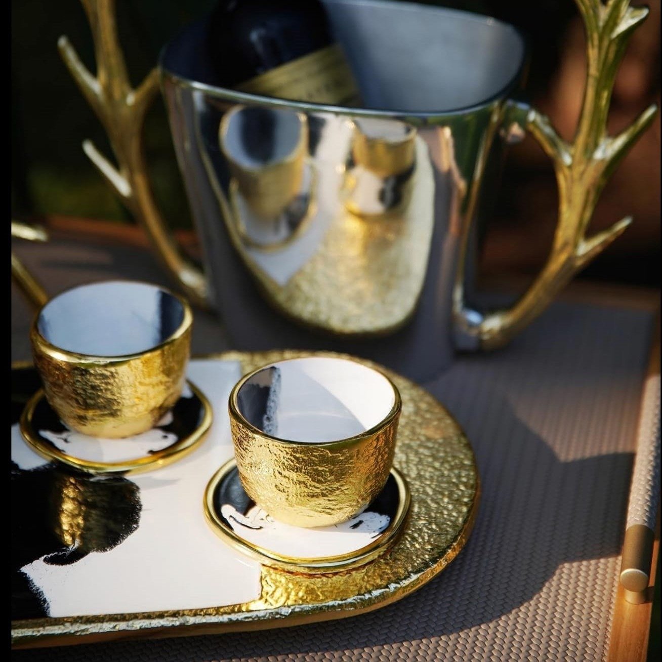 Gold ceramic cups and saucers on a tray with a reflective silver teapot in the background