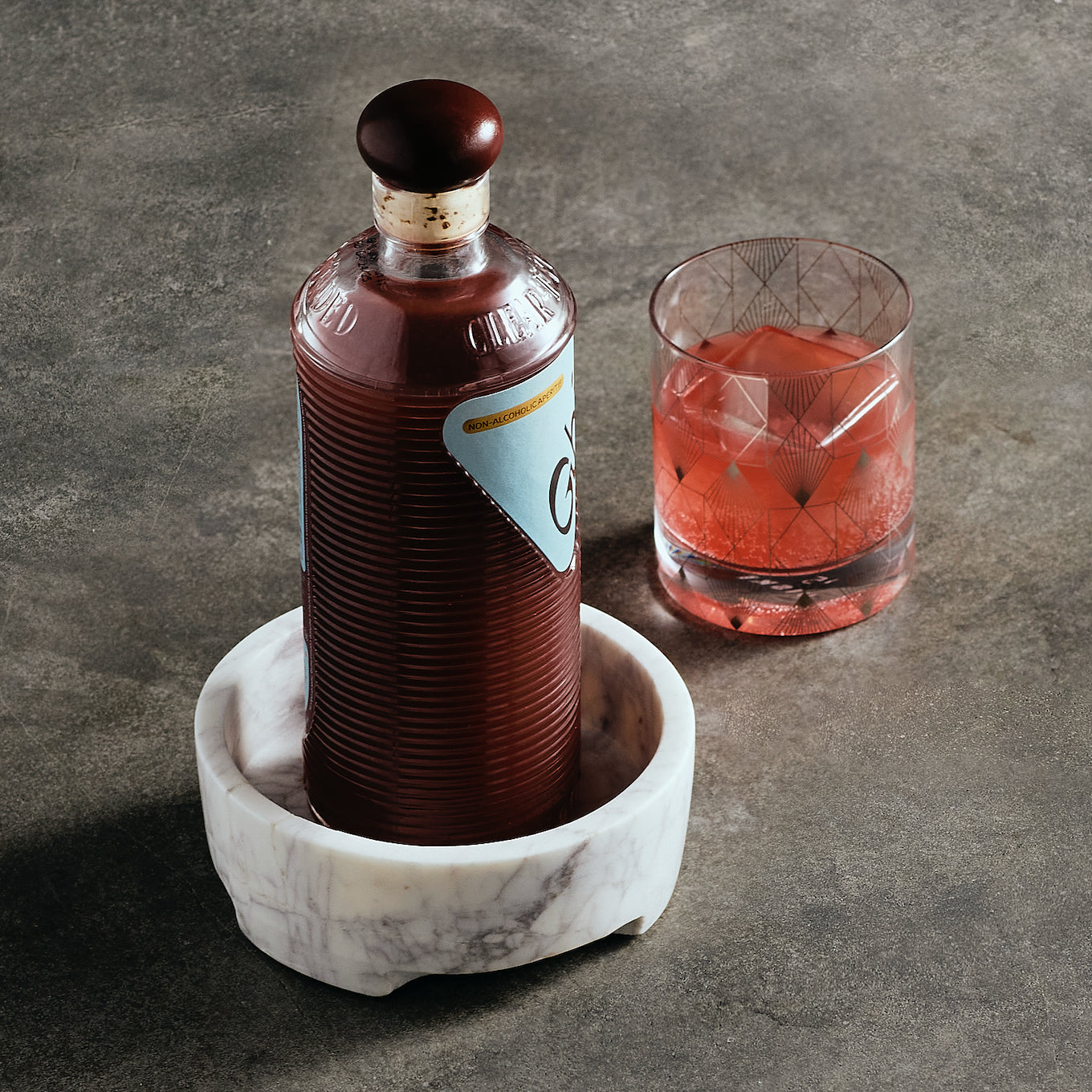 Bottle of red liquid on a marble coaster with a glass of red cocktail in the background