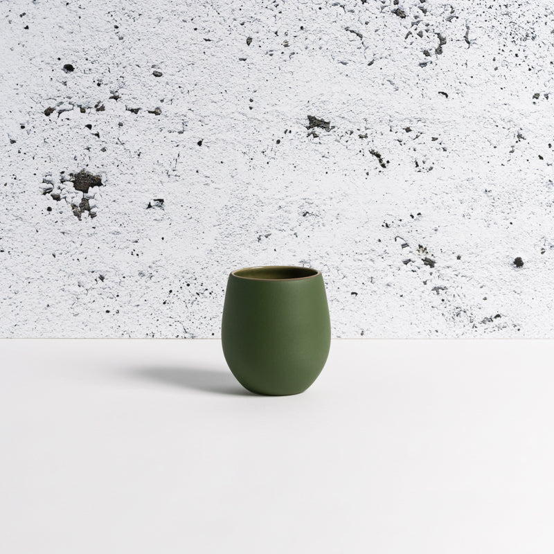 A matte green stoneware coffee cup placed against a textured white background