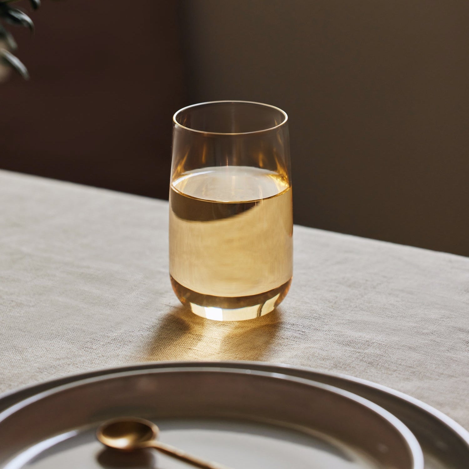 A clear mouth-blown glass cup filled with a golden beverage, placed on a table with a soft lighting environment